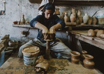 Artisan shaping clay on a potter's wheel in a traditional workshop filled with pottery items.